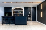 Frontalansicht einer modernen Designküche mit dunkelblauen, geriffelten Fronten, großer Kücheninsel mit zwei Barhockern, dunkler Naturstein-Arbeitsplatte und goldfarbener Armatur, beleuchtet durch Decken-Spotlights und vertikale Lichtleisten