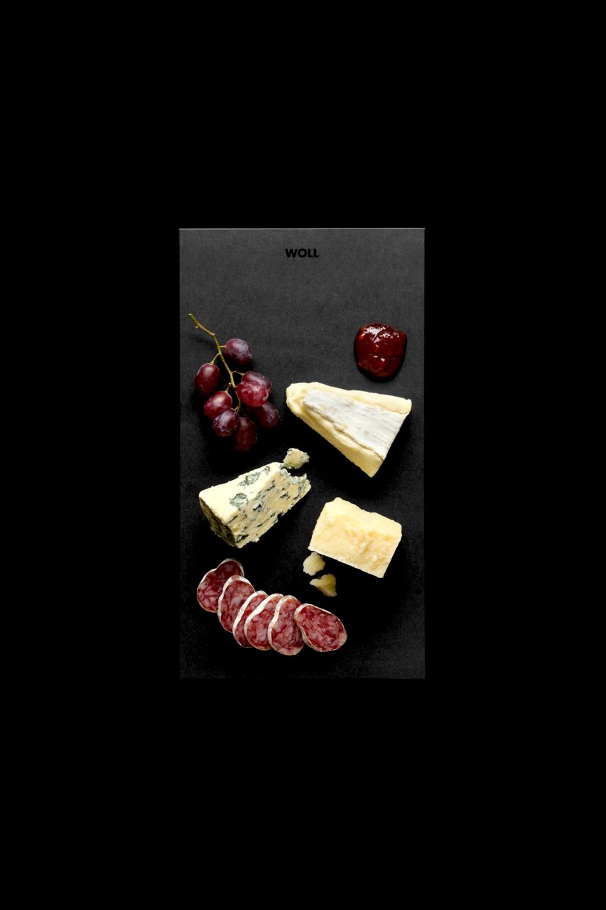 Vogelperspektive auf ein Frühstücksset mit Käse, Wurst, Trauben und Marmelade auf einer schwarzen Platte.