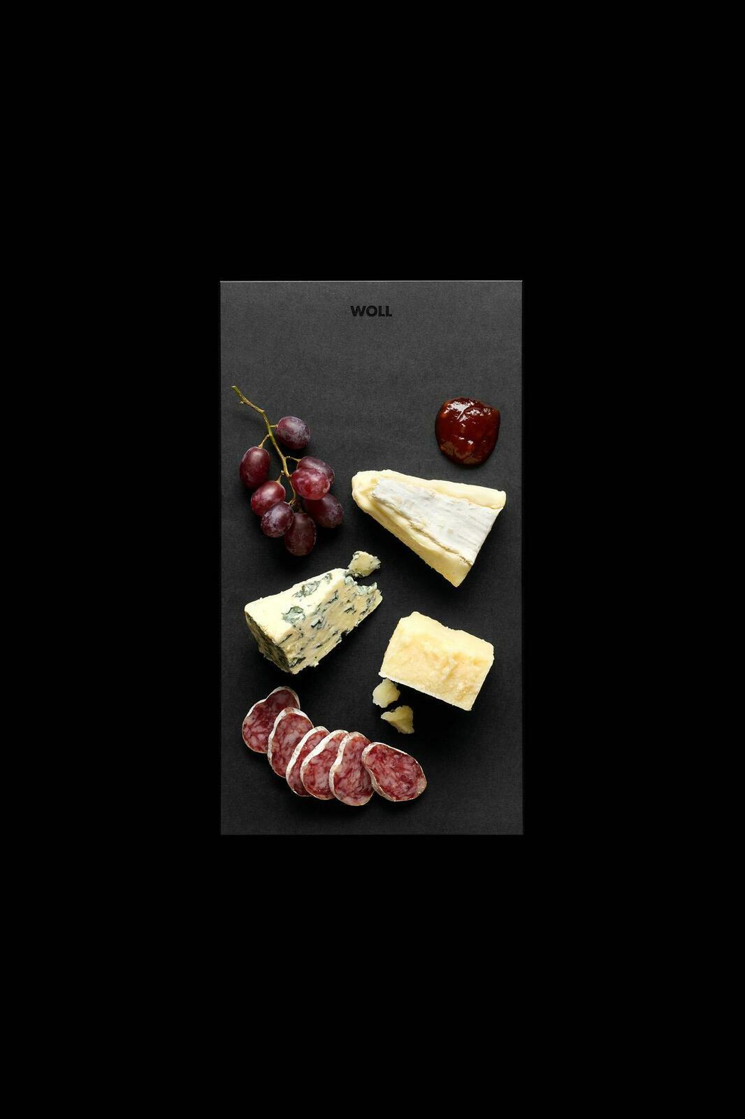 Vogelperspektive auf ein Frühstücksset mit Käse, Wurst, Trauben und Marmelade auf einer schwarzen Platte.