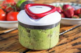 Nahaufnahme eines Easy Pull Universalhackers mit rotem Griff und transparentem Behälter, gefüllt mit Guacamole, auf einem Holztisch. Im Hintergrund sind unscharf Tomaten und Erdbeeren zu sehen.