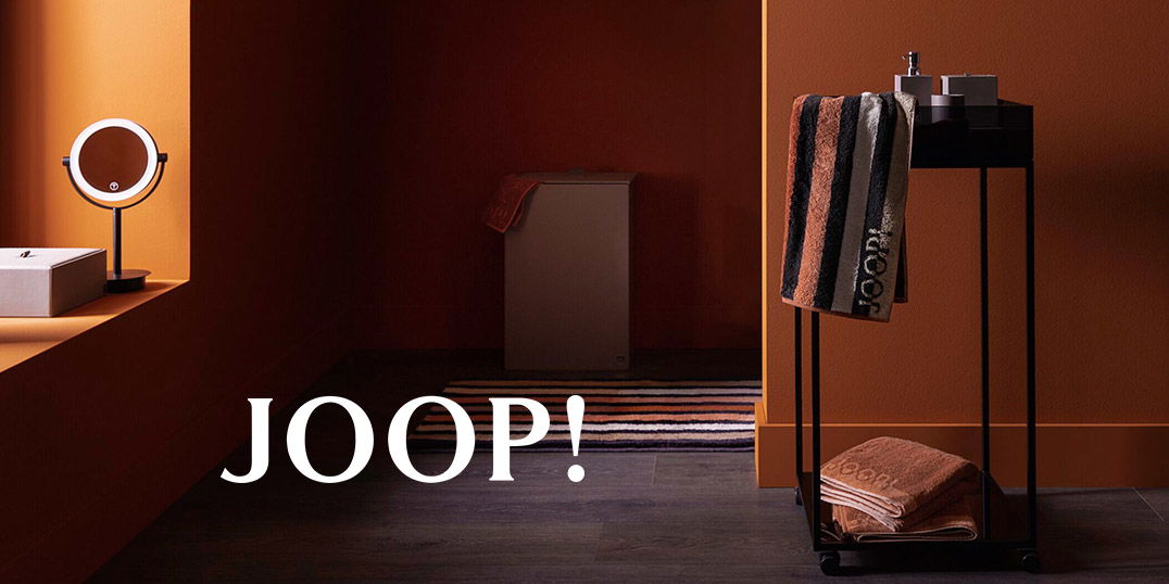 Badezimmer mit einer warmen orangefarbenen Wand, einer modernen Spiegellampe und JOOP! Handtüchern auf einem schwarzen Ständer.