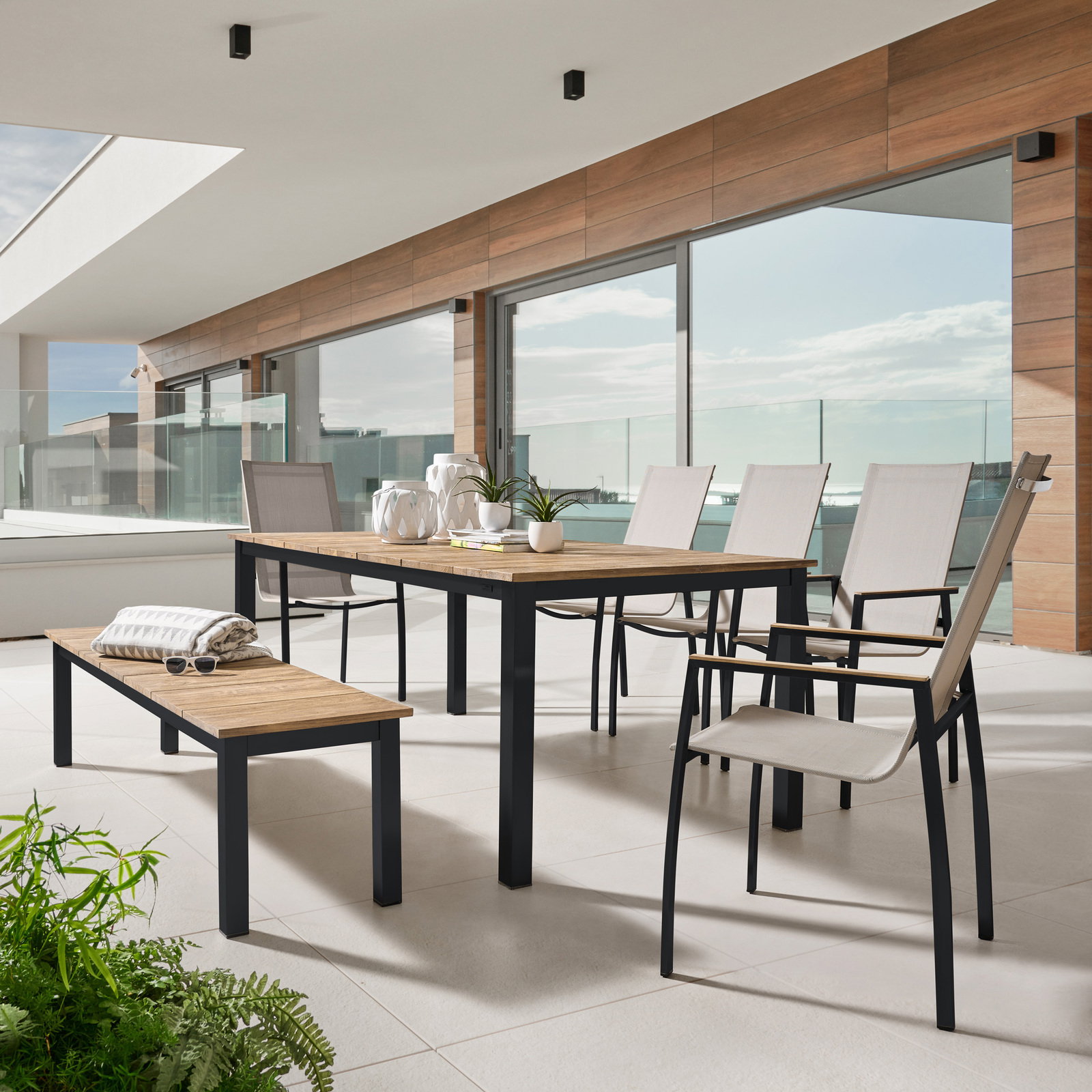 Moderner Gartentisch mit Holzplatte und Metallgestell auf einer Terrasse, umgeben von Stühlen und einer Bank, seitliche Perspektive.