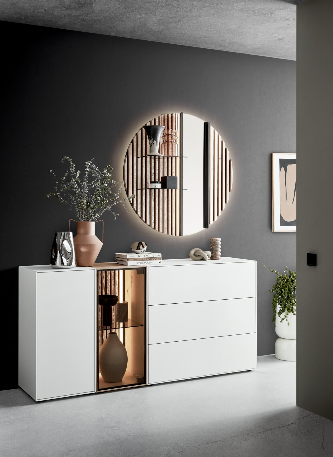 Modernes Sideboard mit Glas links, dekoriert mit Vasen und Büchern, in einem stilvollen Raum mit rundem Spiegel an der Wand. Perspektive von vorne.