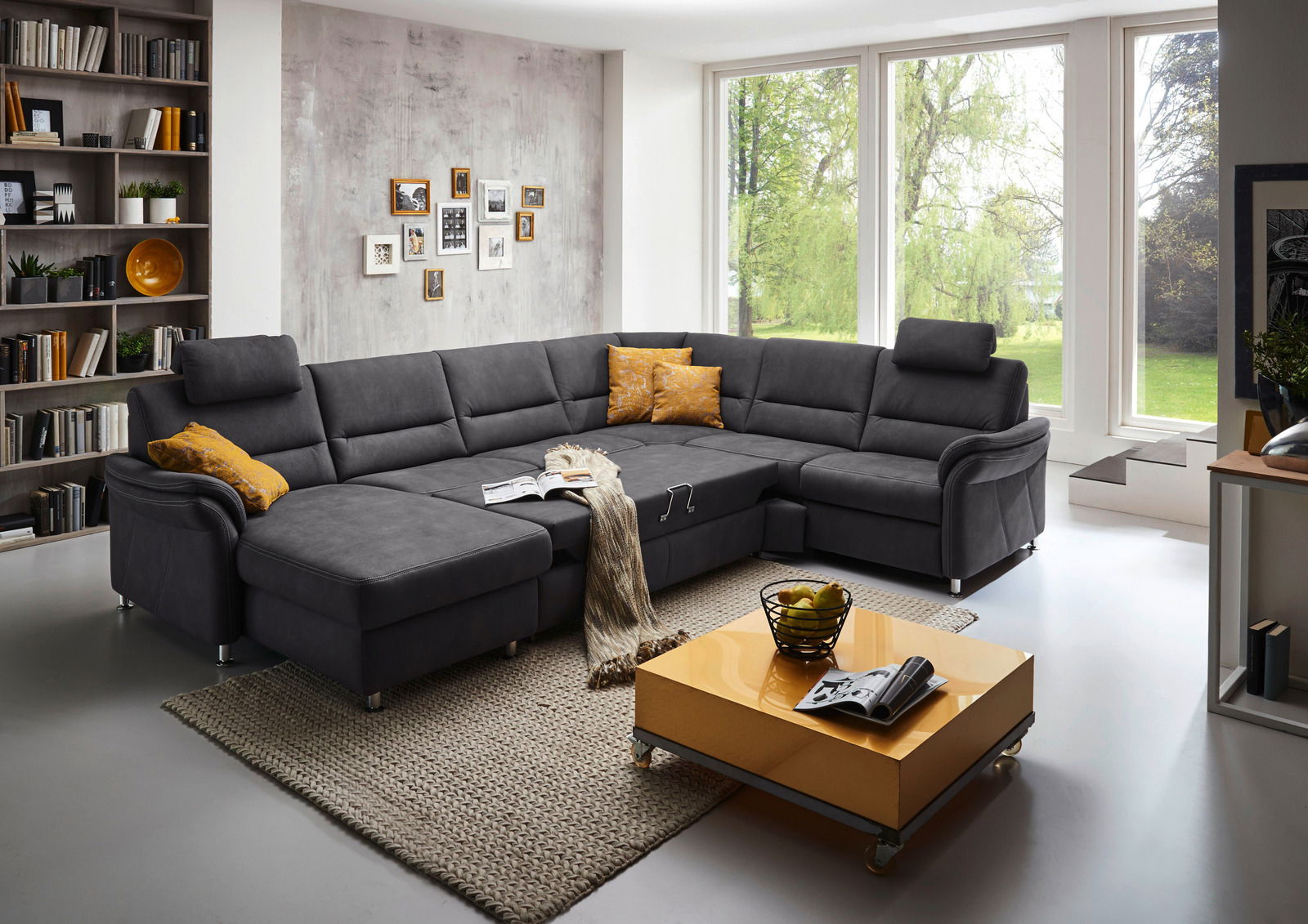 Beldomo System Ecksofa DAKOTA S Moderne Polsterrundecke in dunklem Grau, fotografiert aus einer leicht erhöhten Perspektive in einem hellen Wohnzimmer mit großen Fenstern und stilvoller Dekoration.