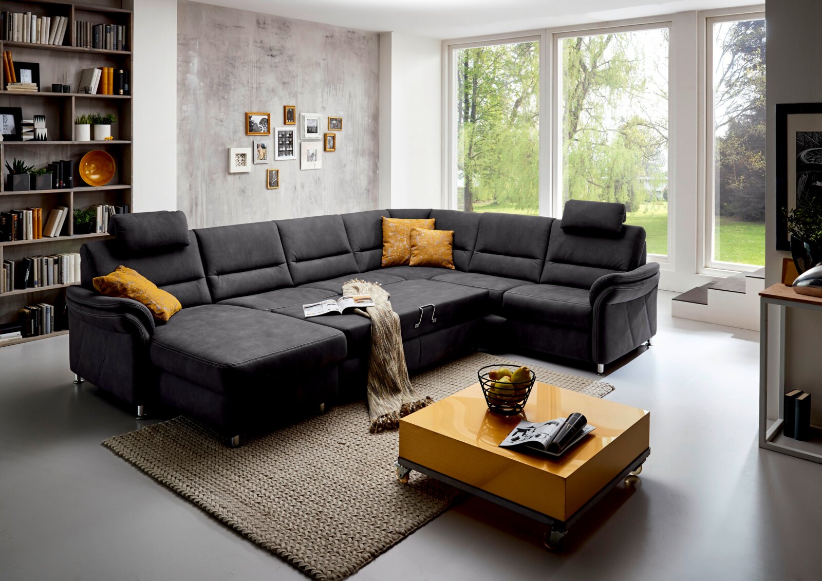 Moderne Polsterrundecke in dunklem Grau, fotografiert aus einer leicht erhöhten Perspektive in einem hellen Wohnzimmer mit großen Fenstern und stilvoller Dekoration.
