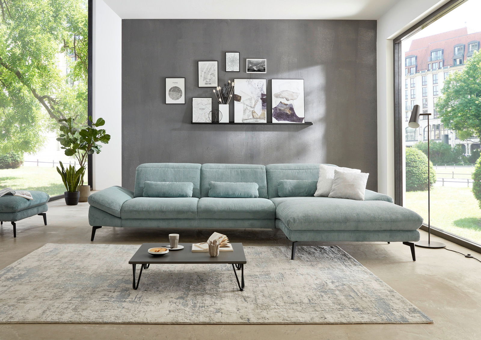 Moderne Longchairkombination in einem hellen Wohnzimmer mit großen Fenstern, grauer Wand und dekorativen Bildern. Perspektive von vorne.