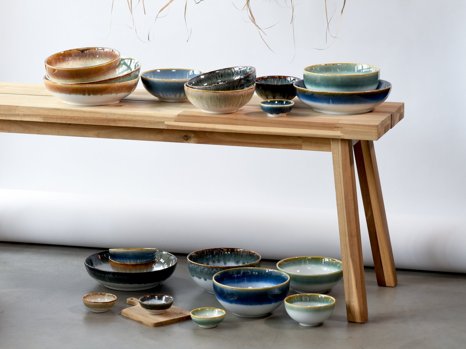 Keramikschalen und kleine Schälchen in Blau-, Grün- und Brauntönen mit reaktiver Glasur auf einem hellen Holztisch arrangiert, zusätzliche Schalen auf dem Boden davor, seitliche Ansicht auf Augenhöhe