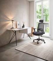 Moderner Schreibtisch mit schlanken Metallbeinen und Schubladen, seitliche Perspektive, neben einem ergonomischen Bürostuhl in einem hellen Raum mit großem Fenster.