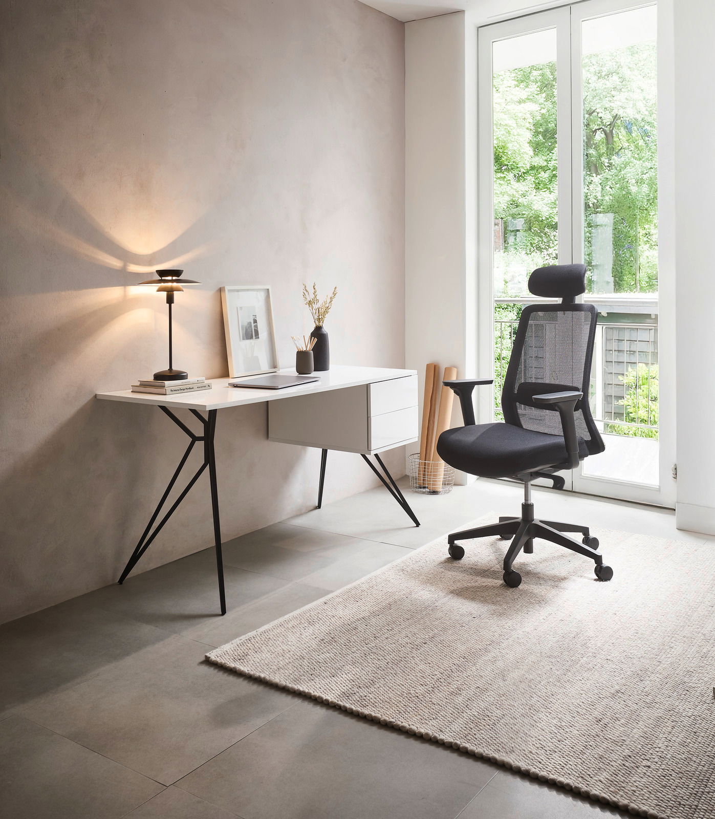 Moderner Schreibtisch mit schlanken Metallbeinen und Schubladen, seitliche Perspektive, neben einem ergonomischen Bürostuhl in einem hellen Raum mit großem Fenster.