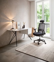 Moderner Schreibtisch mit schlanken Metallbeinen und Schubladen, seitliche Perspektive, neben einem ergonomischen Bürostuhl in einem hellen Raum mit großem Fenster.
