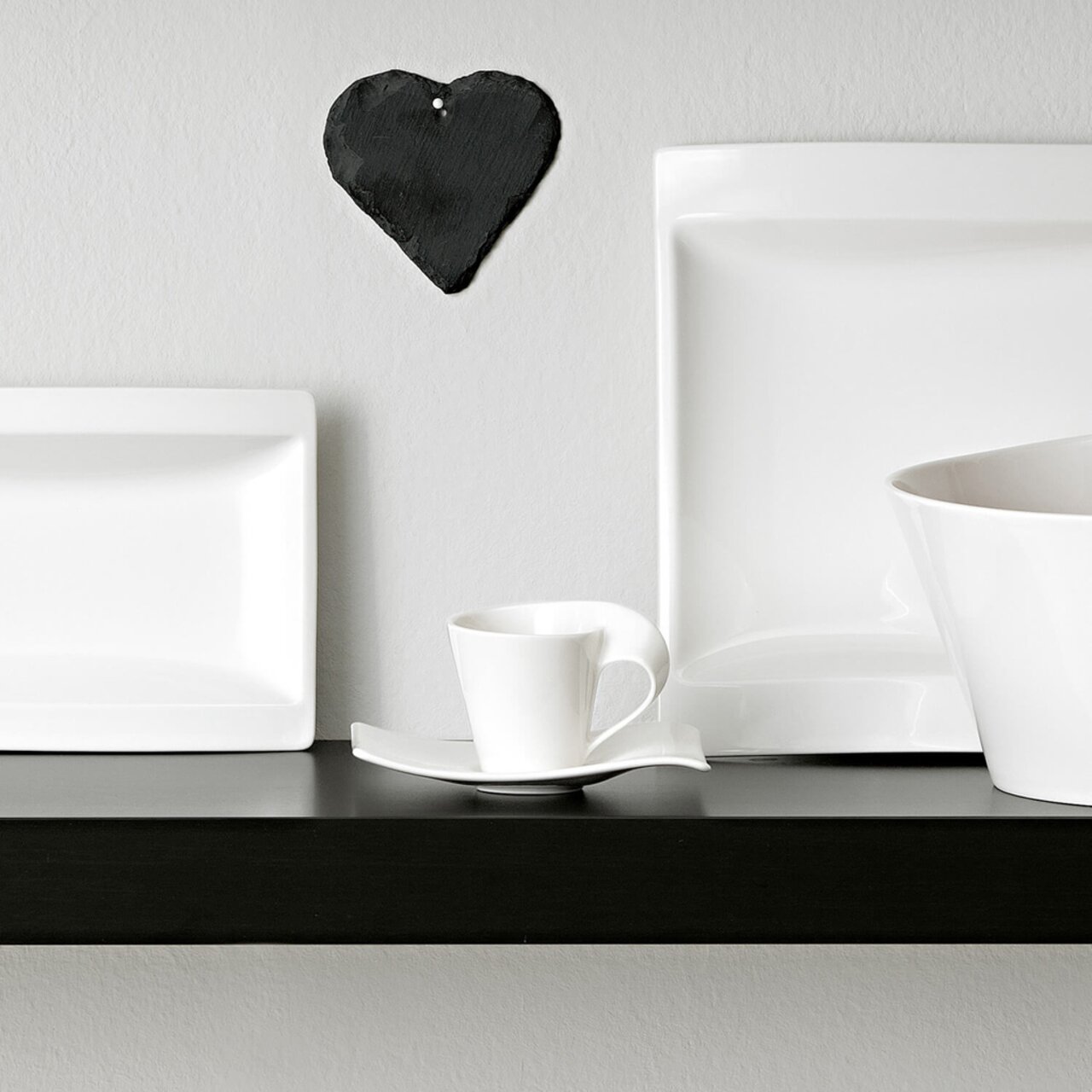 Villeroy & Boch Espresso Untertasse NEW WAVE Weiße Espressotasse mit passender Untertasse auf einem schwarzen Regal, umgeben von modernen, weißen Keramiktellern, aus seitlicher Perspektive fotografiert.