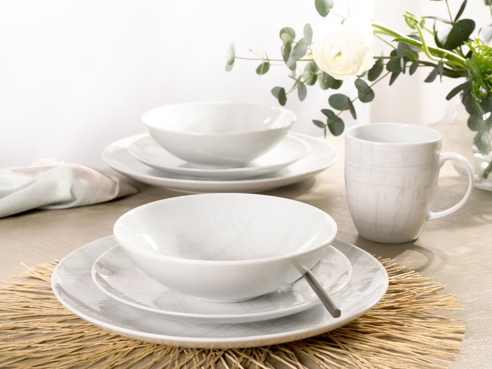 Weißes Porzellan-Geschirrset mit Suppenteller, Speiseteller und Tasse mit dezentem grauen Linienmuster auf gedecktem Tisch, Perspektive: leicht schräg von vorne (3/4-Ansicht).