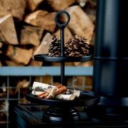 Schwarze zweistöckige Metall-Etagere mit Ringgriff, dekoriert mit Tannenzapfen, Birkenzweigen und gebündelten Zimtstangen, frontal aus leichter Obersicht vor gestapeltem Kaminholz