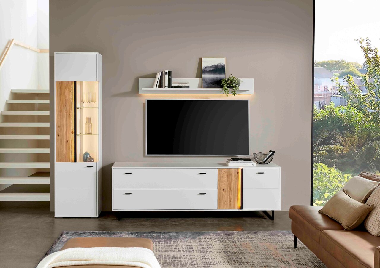 Modern tv-lowboard GLASGOW in wit met houten details, hier van voren afgebeeld, naast een hoge kast en onder een wandrek.