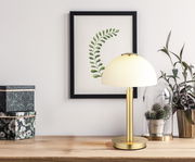 Elegante Tischleuchte mit goldenem Standfuß und weißem Lampenschirm, seitliche Perspektive, auf einem Holztisch neben Pflanzen und dekorativen Boxen.