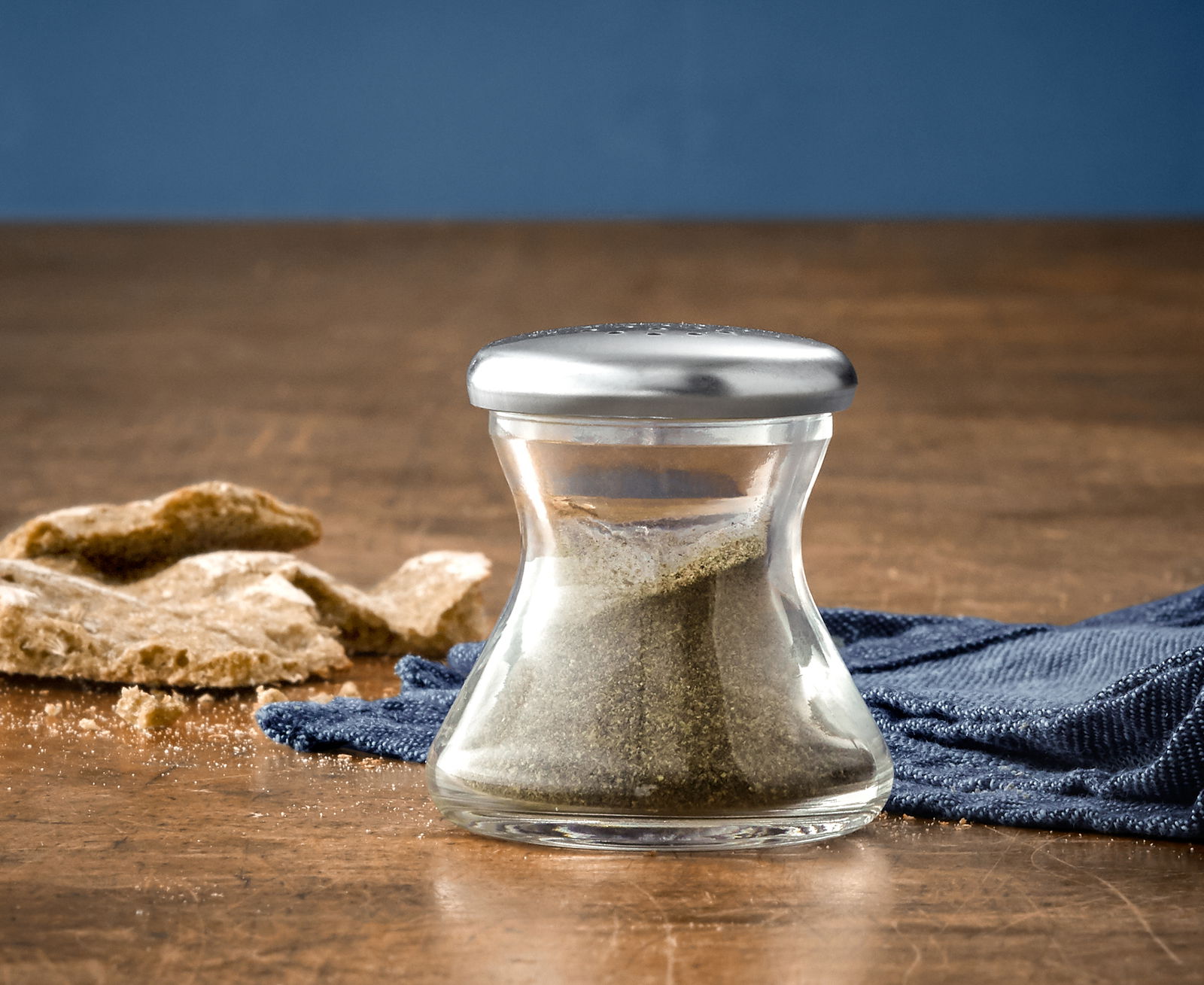 Pfefferstreuer WAGENFELD H 4,5 aus Glas mit silbernem Deckel, gefüllt mit Pfeffer, auf einem Holztisch mit blauer Serviette und Brotkrümeln, seitliche Perspektive.