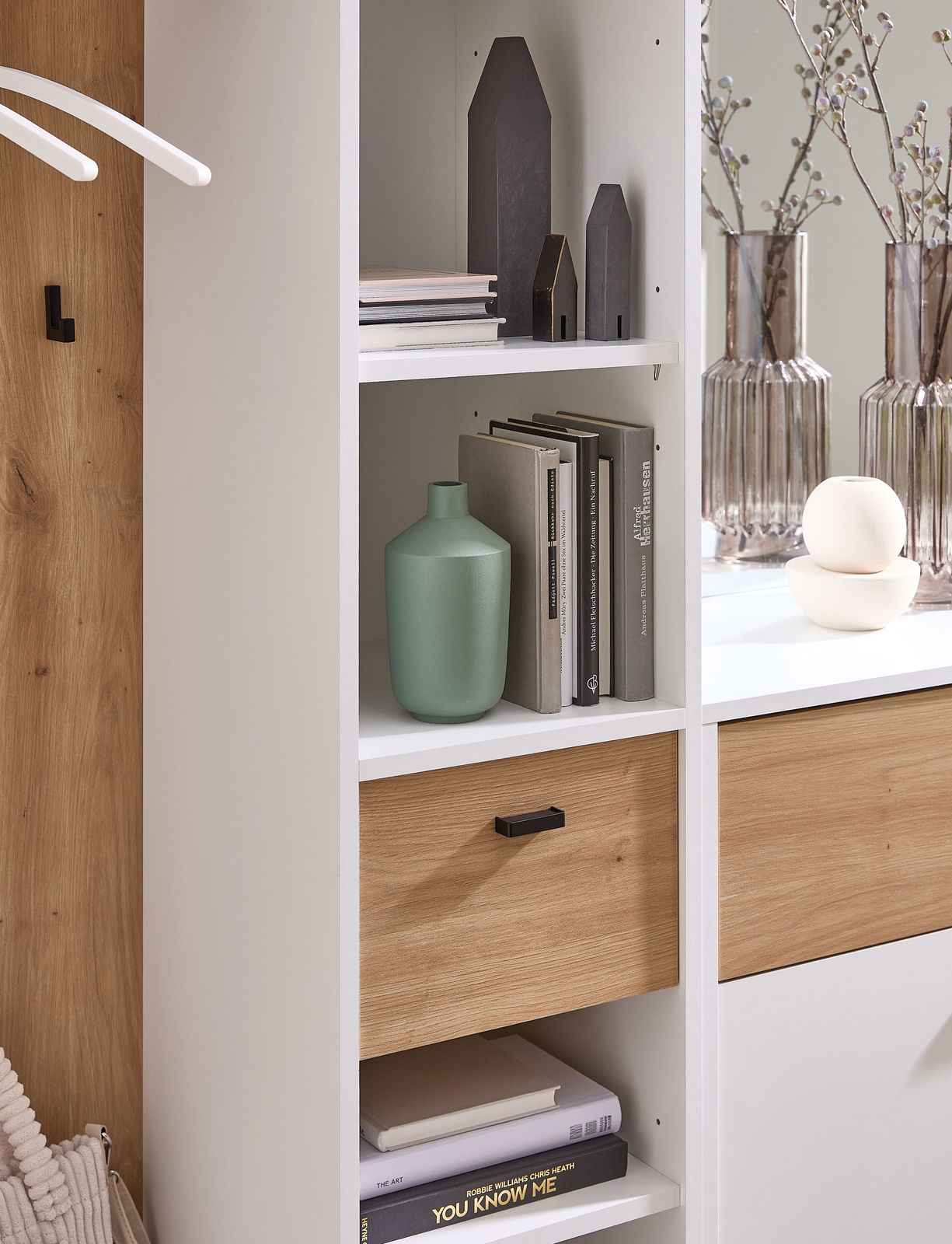 Nahaufnahme einer modernen Kompaktgarderobe mit weißen Regalen und Holzelementen, dekoriert mit Büchern, Vasen und kleinen Deko-Häusern.