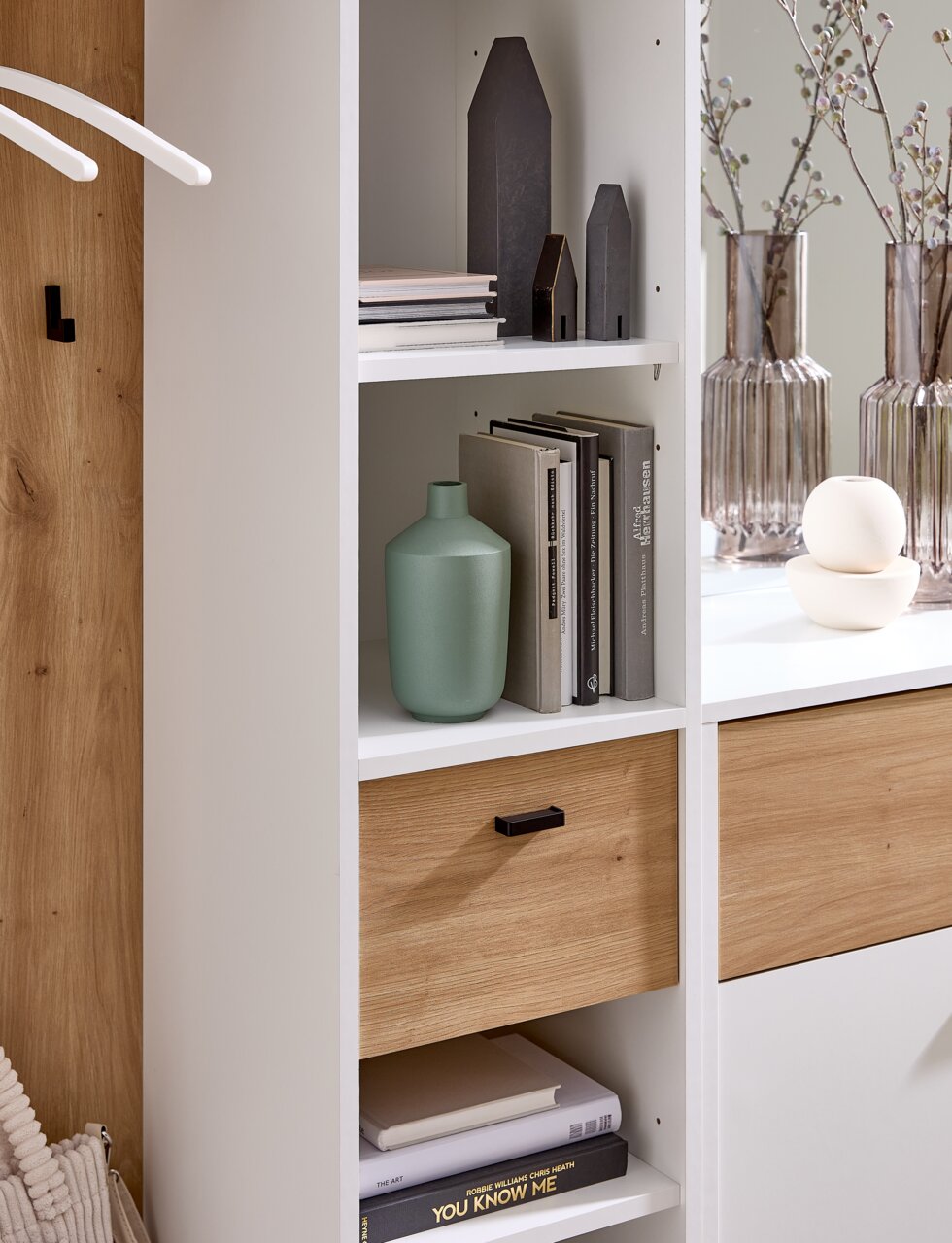 Nahaufnahme einer modernen Kompaktgarderobe mit weißen Regalen und Holzelementen, dekoriert mit Büchern, Vasen und kleinen Deko-Häusern.