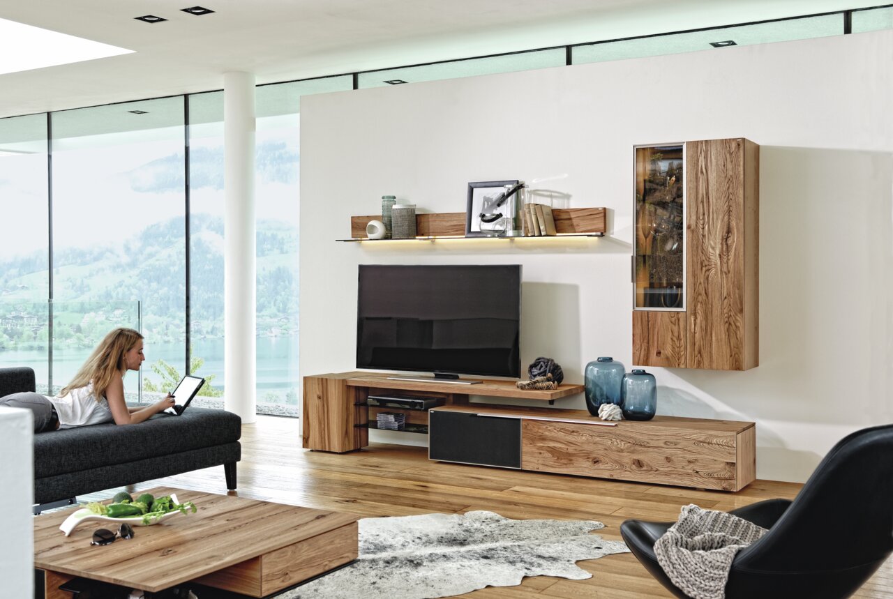 Moderne Wohnwand Voglauer V-ALPIN aus Holz, ohne Beleuchtung, in einem stilvollen Wohnzimmer mit Blick auf die Berge, seitliche Perspektive.