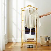 Frontale Ansicht eines hellen Hemdes auf einem Holz-Kleiderständer im minimalistischen Raum, darunter dunkelblaue Shorts, gefaltete Kleidung und graue Hausschuhe; rechts greift eine Hand nach dem Ärmel