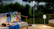 Outdoor-Loungeset auf Terrasse am See mit modernen LED-Steh- und Tischlampen in Schwarz und Weiß, dekorativem bunten Kissen und blauem Plaid, aufgenommen aus leicht erhöhter Frontansicht
