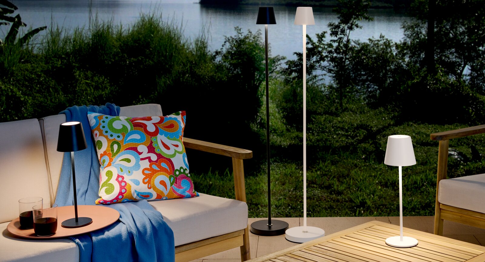 Novel Akku-Tischleuchte SEATTLE Outdoor-Loungeset auf Terrasse am See mit modernen LED-Steh- und Tischlampen in Schwarz und Weiß, dekorativem bunten Kissen und blauem Plaid, aufgenommen aus leicht erhöhter Frontansicht