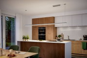 Moderne Küche mit Kücheninsel in Holz- und Weißoptik, integrierten Backöfen und langer LED-Pendelleuchte über der Arbeitsfläche, fotografiert in frontaler Innenraum-Perspektive auf Augenhöhe.