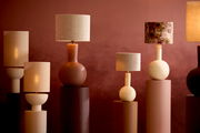 Verschiedene Tischlampen mit runden und zylindrischen Lampenfüßen in warmen Erdtönen, frontal betrachtet vor einer roten Wand.