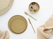 Draufsicht auf ein minimalistisches Tisch-Arrangement mit beigefarbenem Keramikgeschirr (Teller und Schale), goldfarbenem Messer und Gabel sowie einer Schale mit Wachteleiern auf weißem Hintergrund mit hellem Stofftuch.