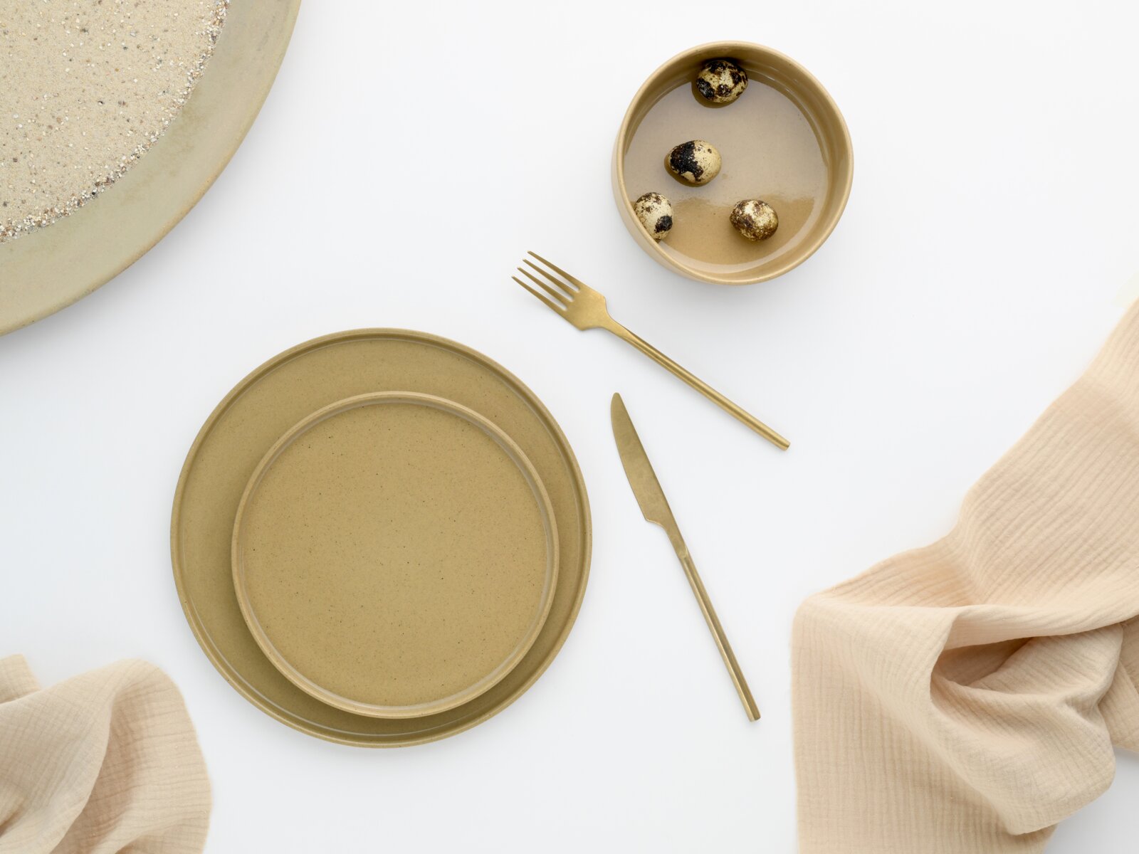 Draufsicht auf ein minimalistisches Tisch-Arrangement mit beigefarbenem Keramikgeschirr (Teller und Schale), goldfarbenem Messer und Gabel sowie einer Schale mit Wachteleiern auf weißem Hintergrund mit hellem Stofftuch.