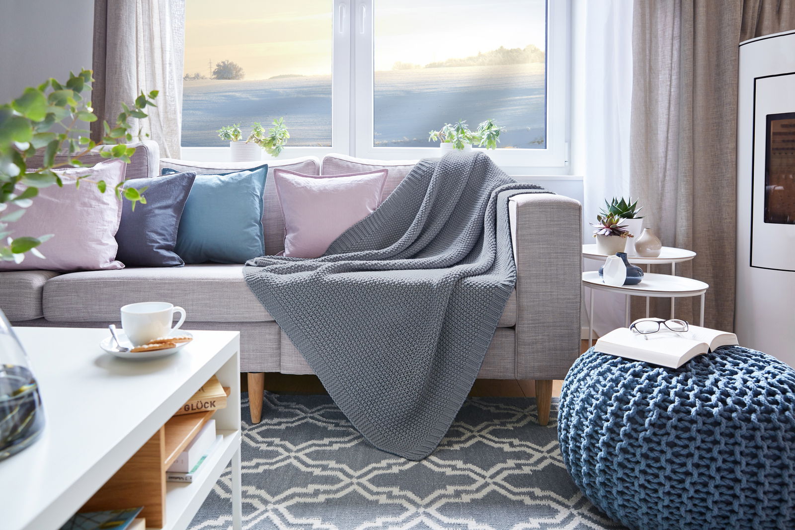 Zurbrüggen Decke Gemütliches Wohnzimmer mit grauer Strickdecke auf einem Sofa, umgeben von pastellfarbenen Kissen, mit Blick aus dem Fenster auf eine winterliche Landschaft.