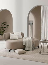 Eleganter Wohnraum mit einem beigen Sofa, dekoriert mit Kissen und einer Decke von JOOP!, im Vordergrund ein runder Beistelltisch mit Tasse und Vase, im Hintergrund ein Vorhang in einem modernen, hellen Raum mit Pflanzen und Bögen.