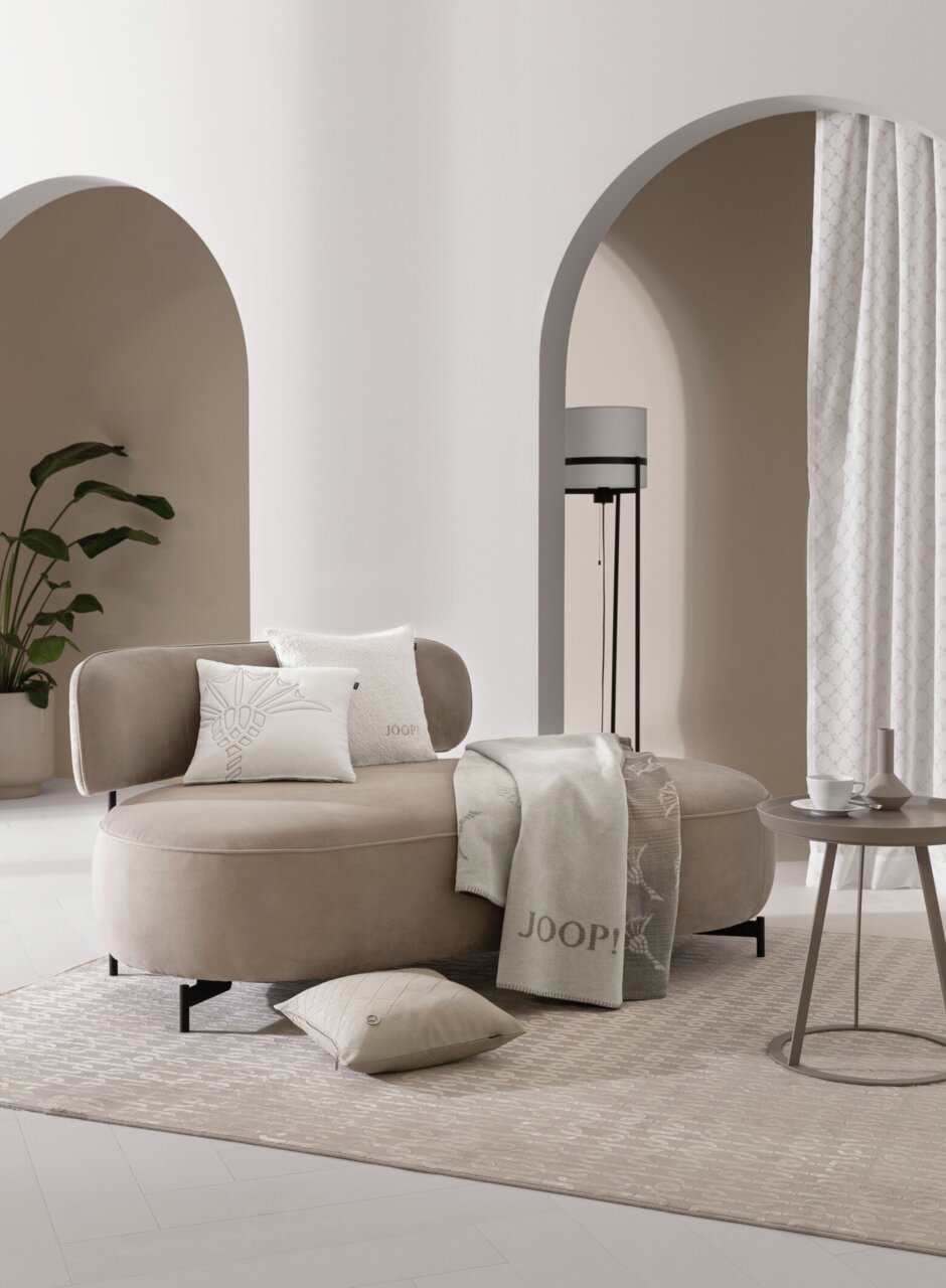 Eleganter Wohnraum mit einem beigen Sofa, dekoriert mit Kissen und einer Decke von JOOP!, im Vordergrund ein runder Beistelltisch mit Tasse und Vase, im Hintergrund ein Vorhang in einem modernen, hellen Raum mit Pflanzen und Bögen.