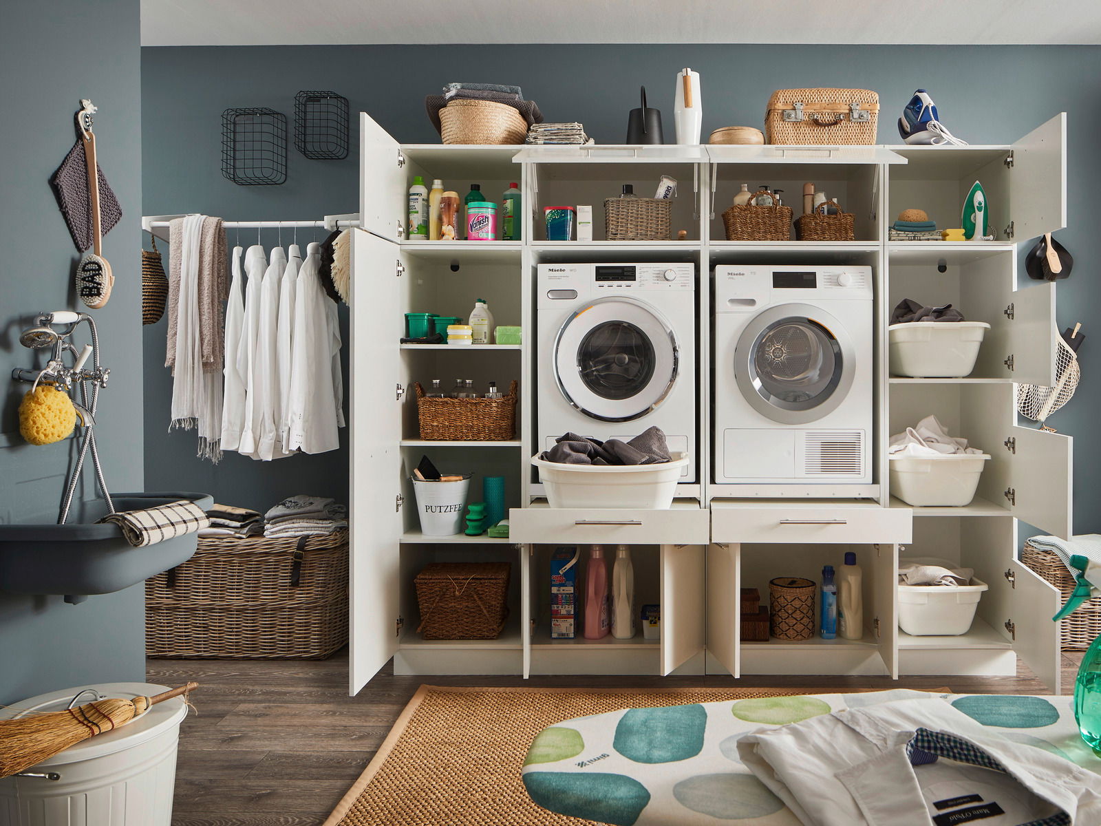 Vorderansicht eines modernen Hauswirtschaftsraums mit geöffneten Schränken, die Waschmaschine, Trockner und verschiedene Haushaltsgegenstände zeigen.