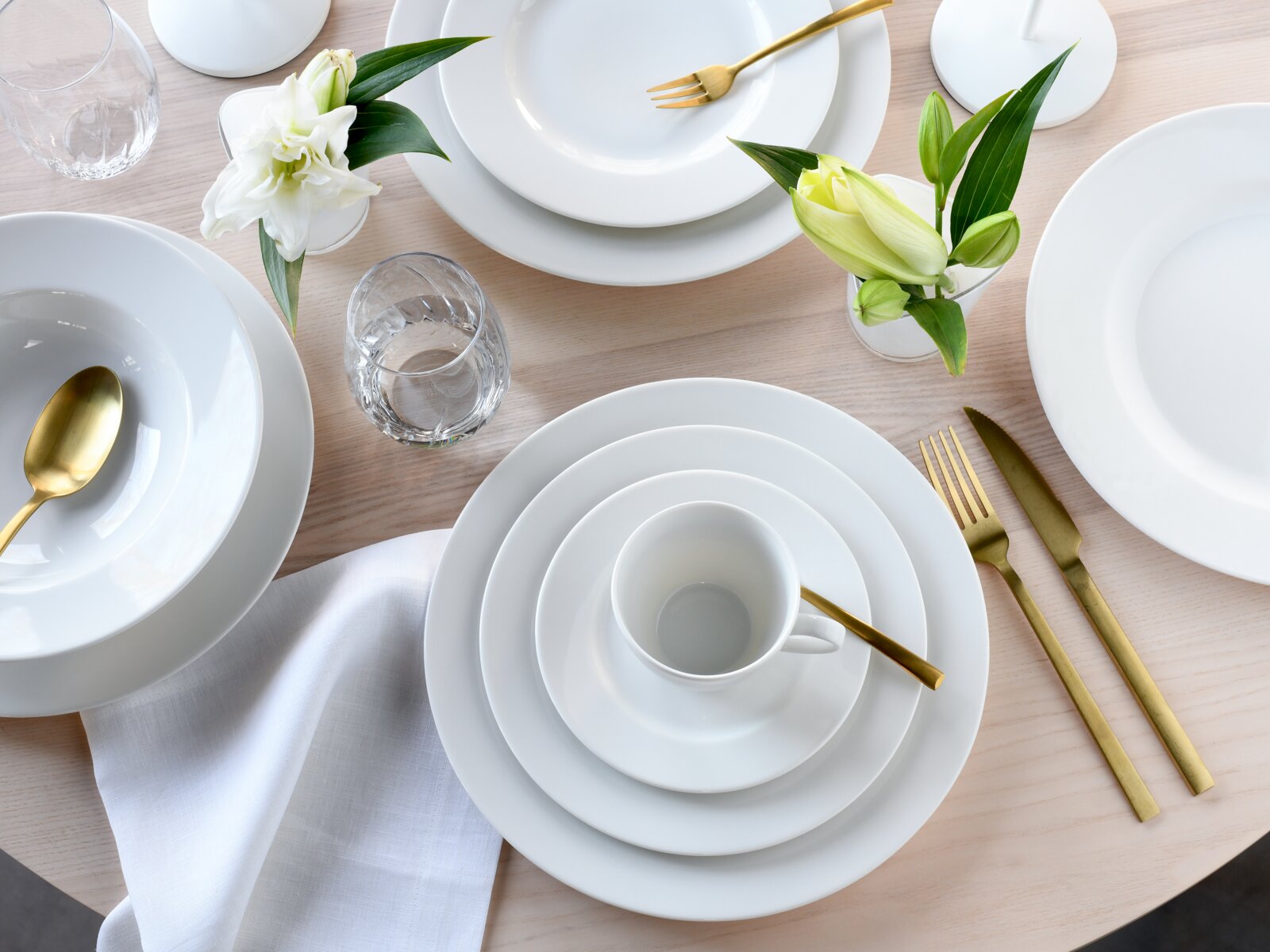 Draufsicht auf modern gedeckten Esstisch mit weißem Porzellan-Geschirr (Teller, Schalen und Tasse), goldfarbenem Besteck, Wassergläsern und Lilien in kleinen Vasen auf hellem Holztisch