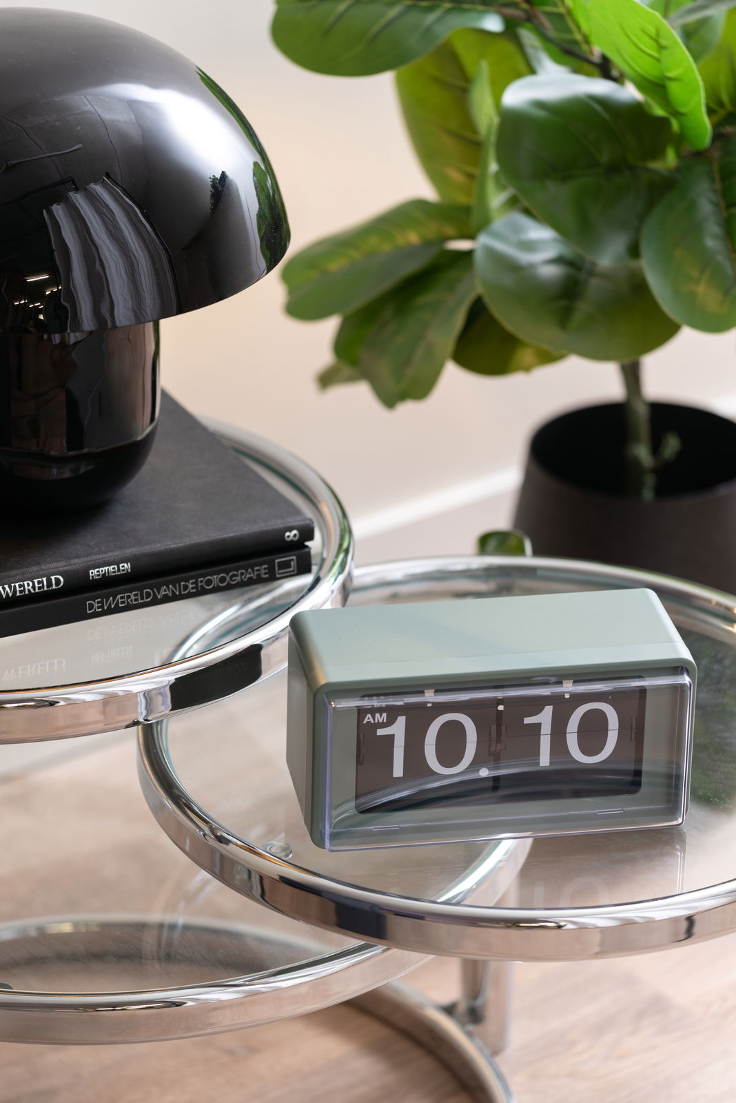 Grüne Flip-Uhr auf einem runden, verchromten Tisch, neben einer schwarzen Lampe und einer Pflanze, aus seitlicher Perspektive fotografiert.