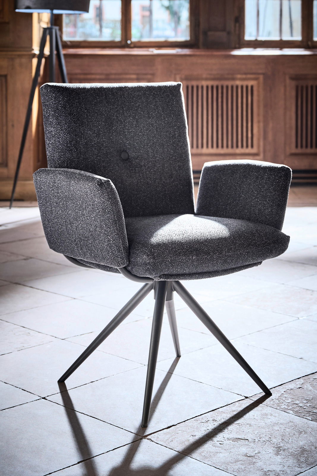 Grauer Armlehnstuhl mit modernem Design, fotografiert aus einem leicht seitlichen Winkel in einem Raum mit Holzwänden und großen Fenstern.