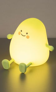 Frontalansicht einer Akku-Nachtleuchte in Form einer gelben, lächelnden Figur mit Armen und Beinen, die auf einem Tisch sitzt.