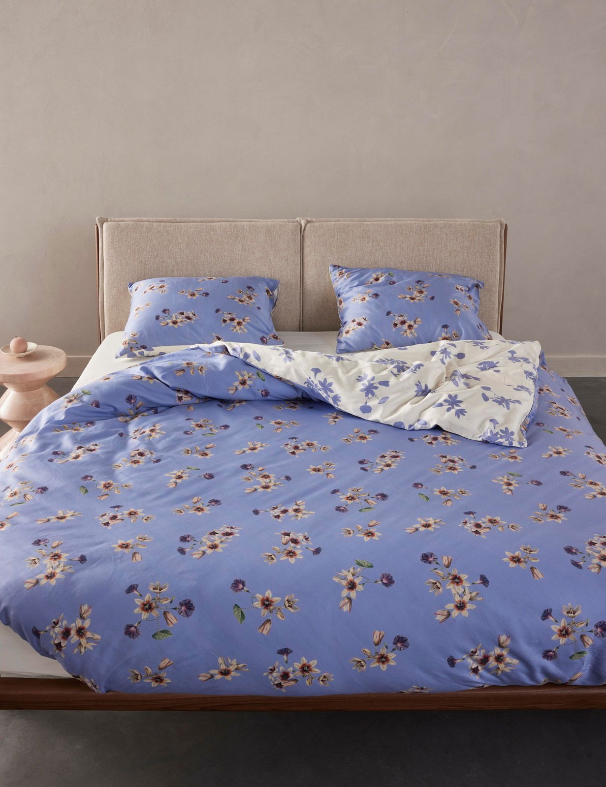 Bettwäsche-Set in Blau mit floralem Muster, bestehend aus Decke und zwei Kissen, auf einem Bett mit beigem Kopfteil, aus der Frontalperspektive fotografiert.