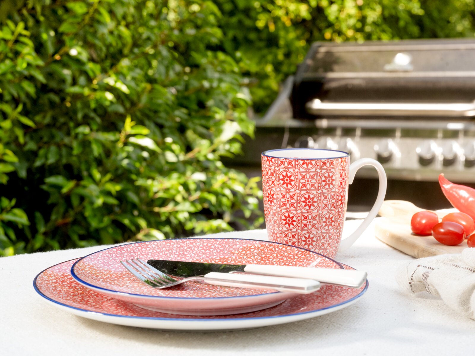 Rot-weiß gemusterter Keramikbecher mit blauem Rand und passendem Teller auf weißer Tischdecke im Garten, seitliche Perspektive mit leichtem Blick von vorn; im Hintergrund ein Grill sowie Tomaten und Chili auf einem Holzbrett.