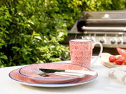 Rot-weiß gemusterter Keramikbecher mit blauem Rand und passendem Teller auf weißer Tischdecke im Garten, seitliche Perspektive mit leichtem Blick von vorn; im Hintergrund ein Grill sowie Tomaten und Chili auf einem Holzbrett.