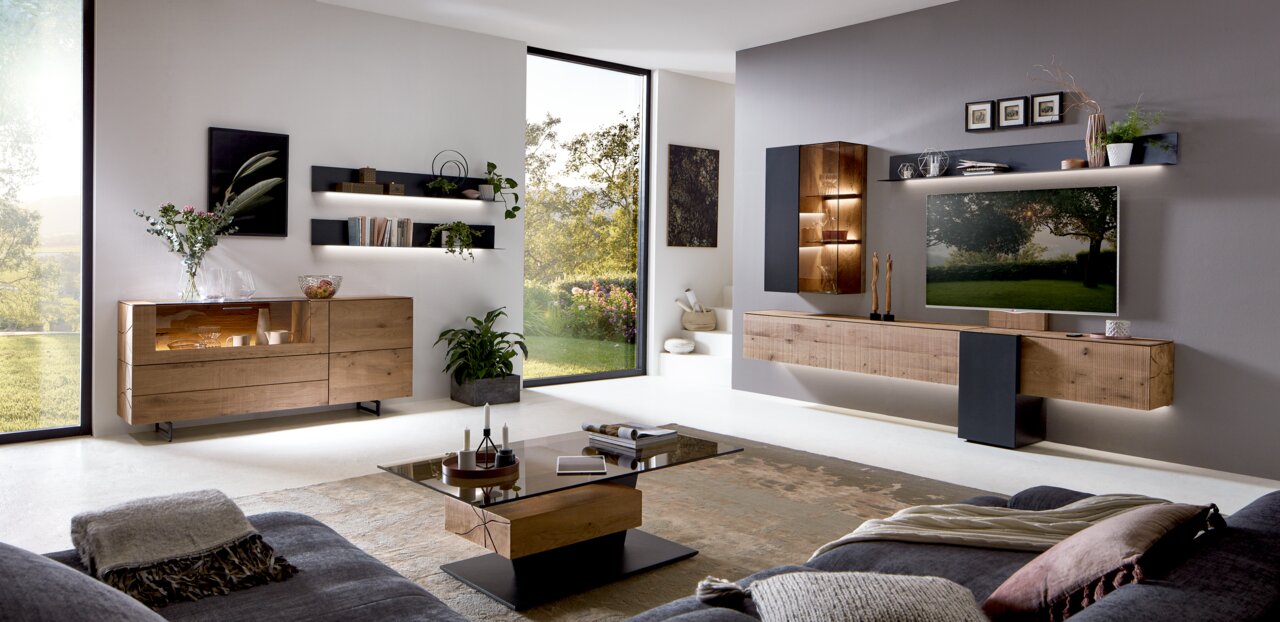 Valnatura Dressoir LOVIS Een moderne woonkamer met een elegant houten dressoir, hier gezien vanaf de zijkant. Het dressoir heeft een glazen vitrinekast en maakt deel uit van een stijlvolle inrichting met wandplanken en een groot raam met uitzicht op het groen.