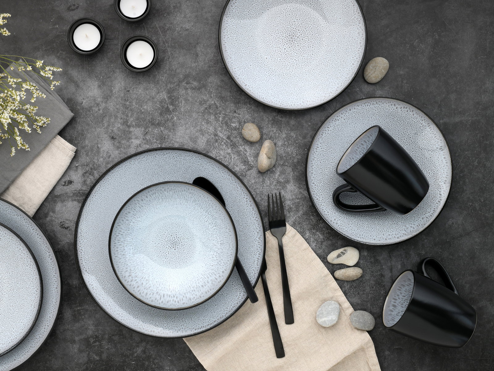 Vogelperspektive auf ein stilvolles 16-teiliges Kombiservice-Set mit grauen Tellern und Schalen sowie schwarzen Tassen auf einem dunklen Untergrund, dekoriert mit Steinen und Kerzen.