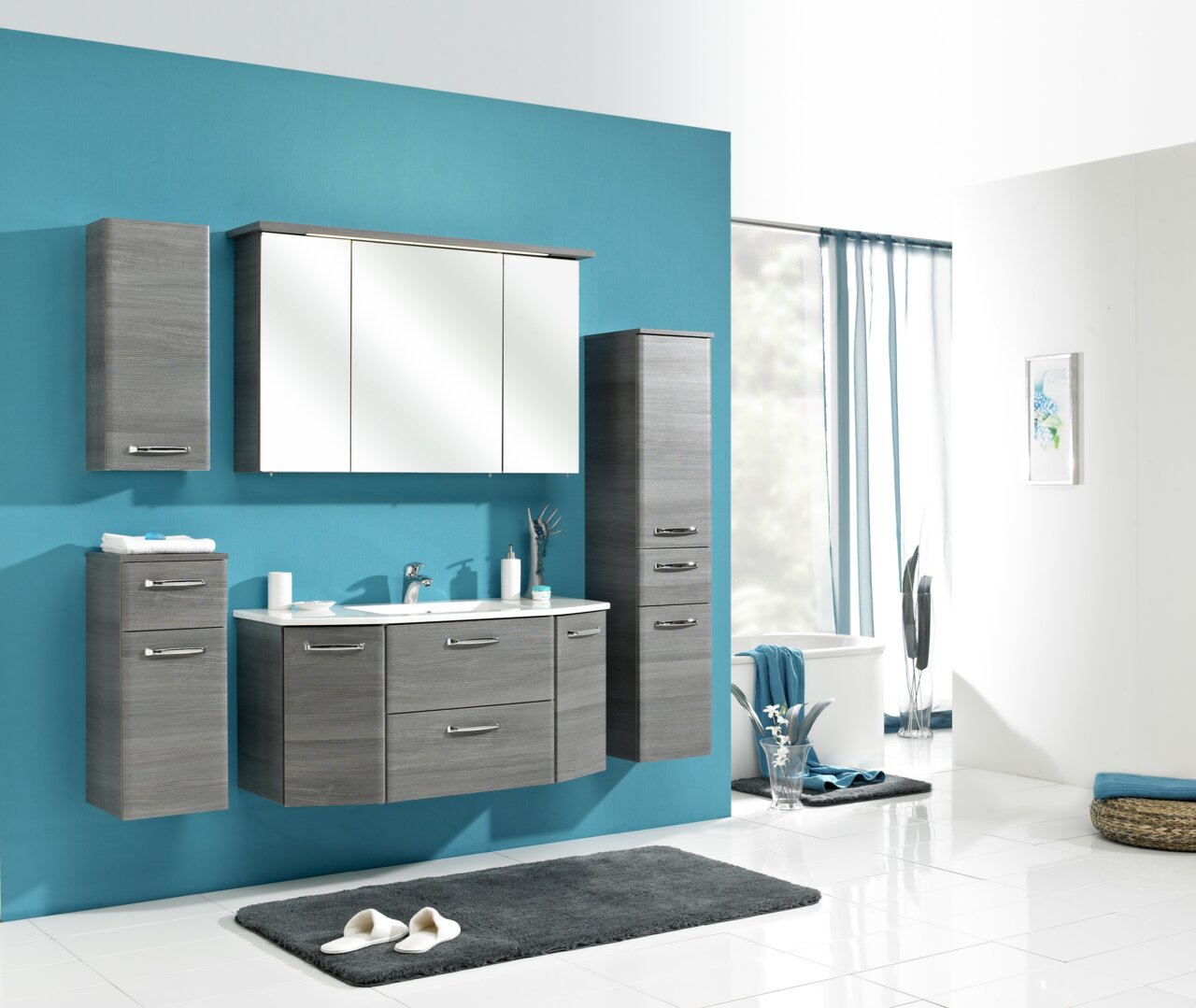 Modernes Badmöbel-Set in grauer Holzoptik mit Waschtisch-Unterschrank, Spiegelschrank und zwei Hochschränken an türkisfarbener Wand, frontal leicht schräg von vorn fotografiert.