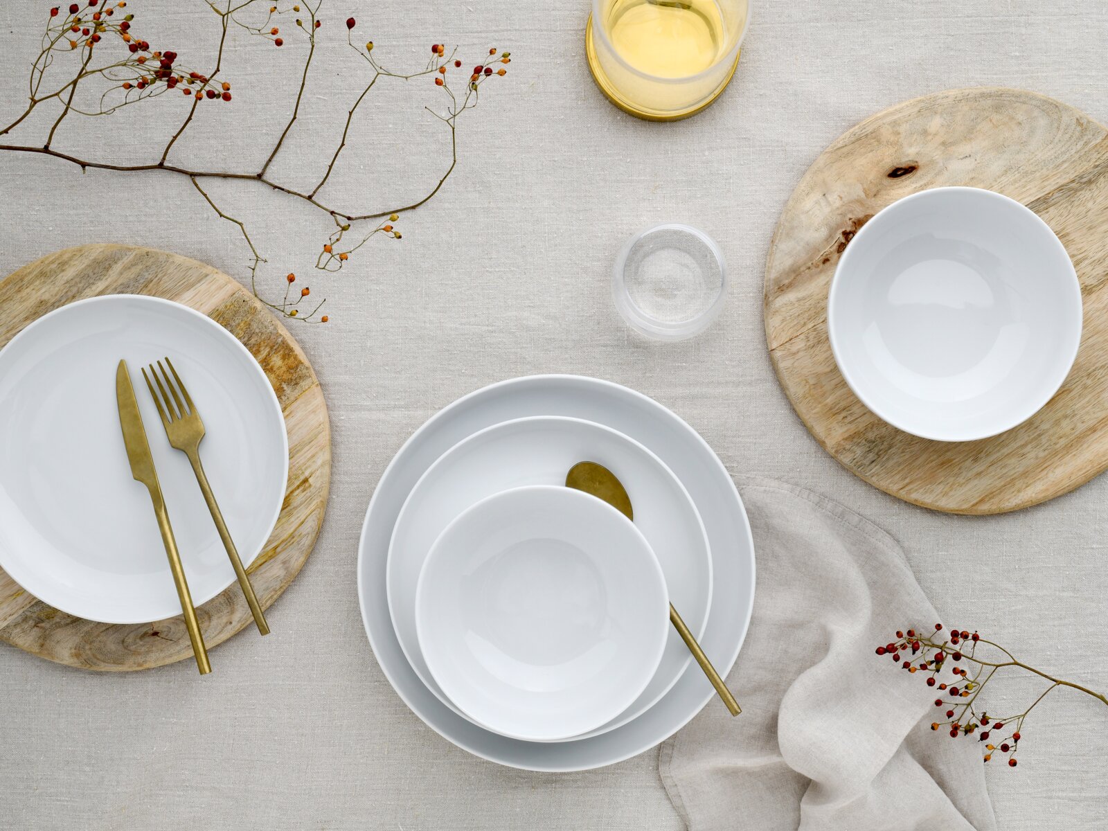 Draufsicht auf ein minimalistisches Tischgedeck mit weißen Porzellantellern und -schalen auf Holzuntersetzern, goldfarbenem Besteck, Leinentischdecke, Glas und dekorativen Beerenzweigen