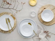 Draufsicht auf ein minimalistisches Tischgedeck mit weißen Porzellantellern und -schalen auf Holzuntersetzern, goldfarbenem Besteck, Leinentischdecke, Glas und dekorativen Beerenzweigen
