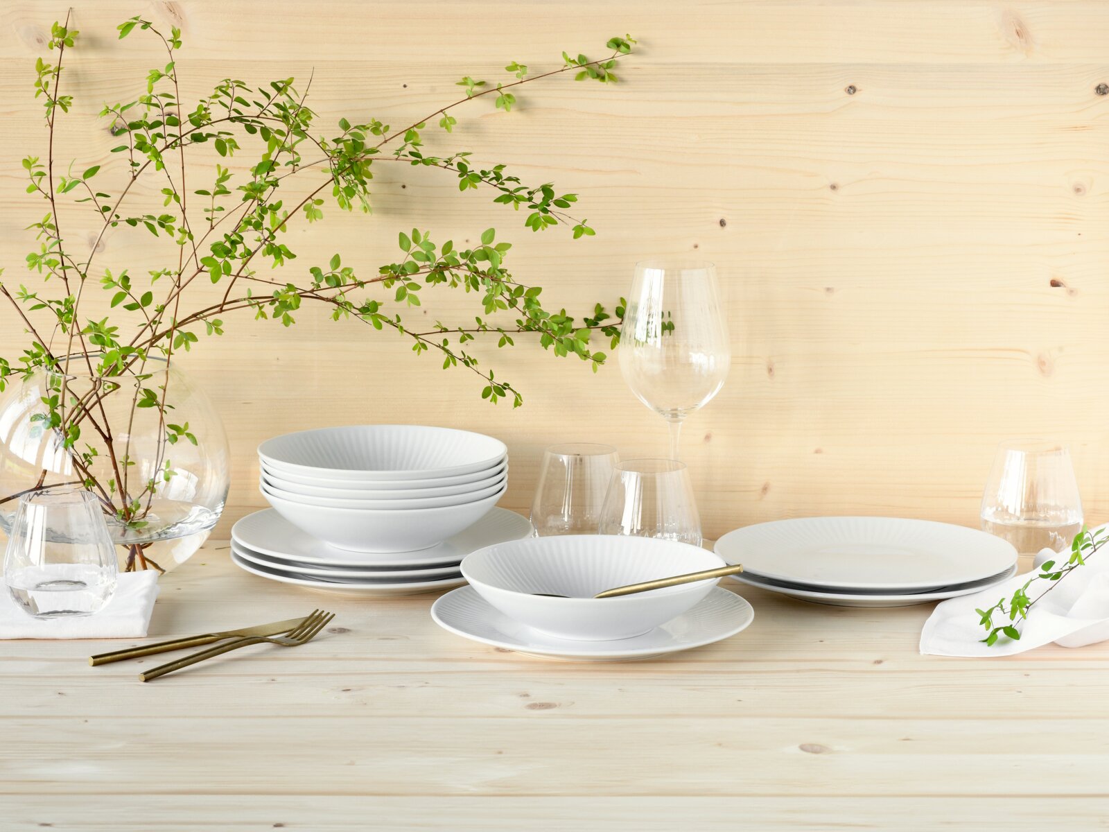 Creatable Tafelservice 12-tlg VALENCIA Minimalistisches weißes Geschirr-Set mit Tellern und Schalen sowie klaren Gläsern und goldfarbenem Besteck auf hellem Holztisch vor Holzpaneel-Wand, dekoriert mit grünen Zweigen, Perspektive: frontale leicht erhöhte Ansicht