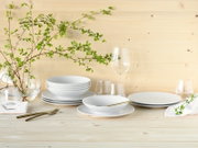 Minimalistisches weißes Geschirr-Set mit Tellern und Schalen sowie klaren Gläsern und goldfarbenem Besteck auf hellem Holztisch vor Holzpaneel-Wand, dekoriert mit grünen Zweigen, Perspektive: frontale leicht erhöhte Ansicht