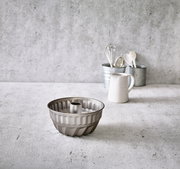 Gugelhupfform aus Metall, frontal auf einem Küchentisch platziert, mit Küchenutensilien im Hintergrund.