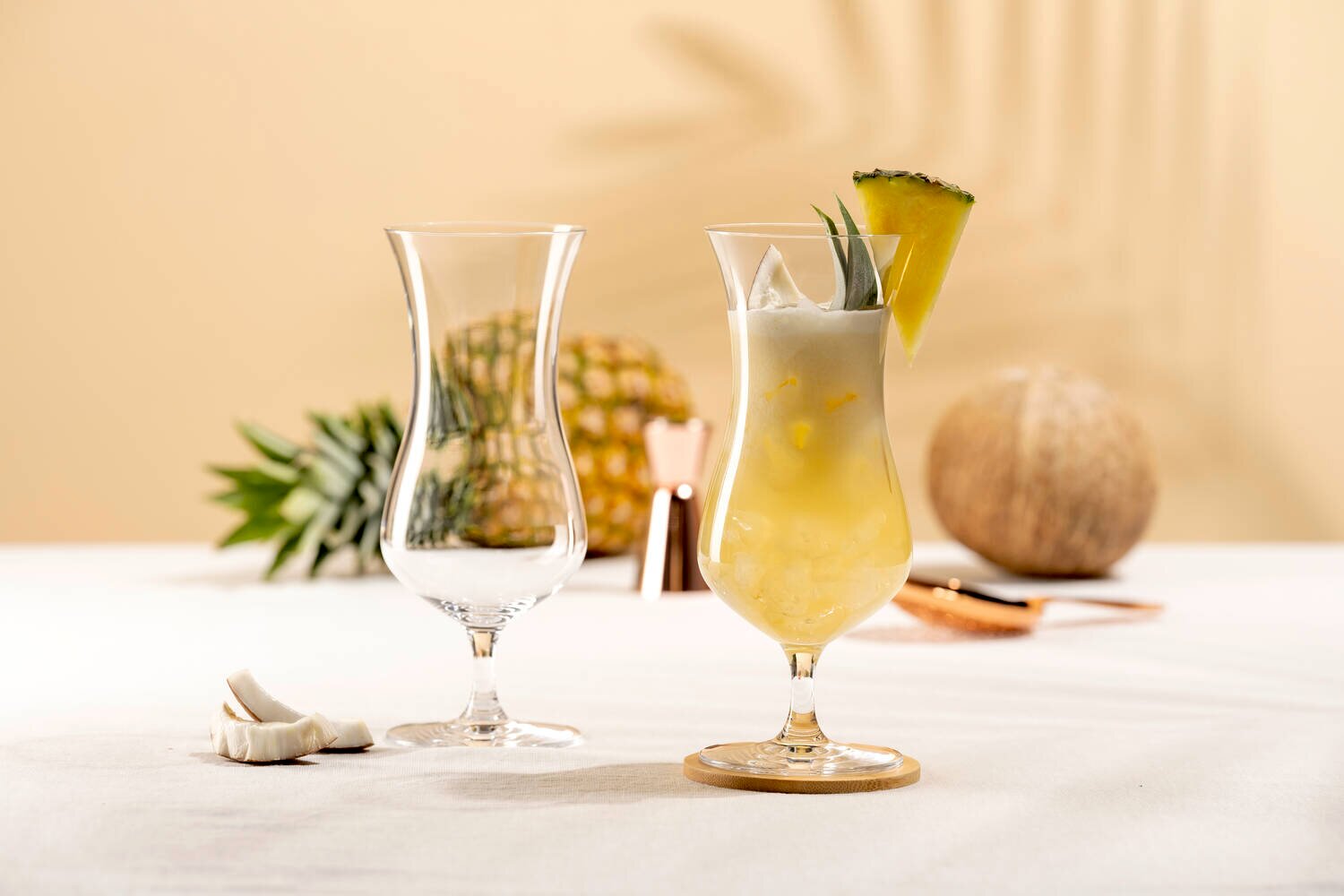 Vooraanzicht van twee elegante hurricane cocktailglazen, één leeg en één gevuld met een romige tropische ananas-kokosdrank, ijs en een schijfje ananas